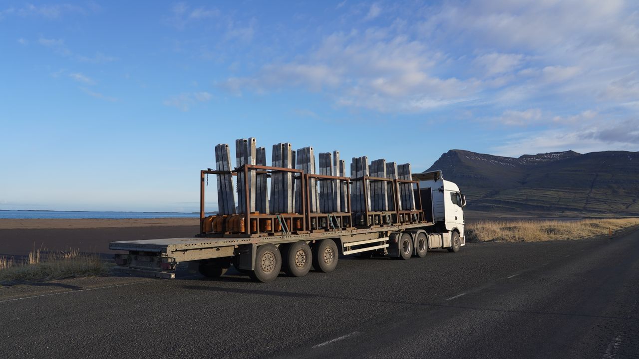 A truck carrying metal cargo on a scenic Icelandic road with mountains in the backdrop.