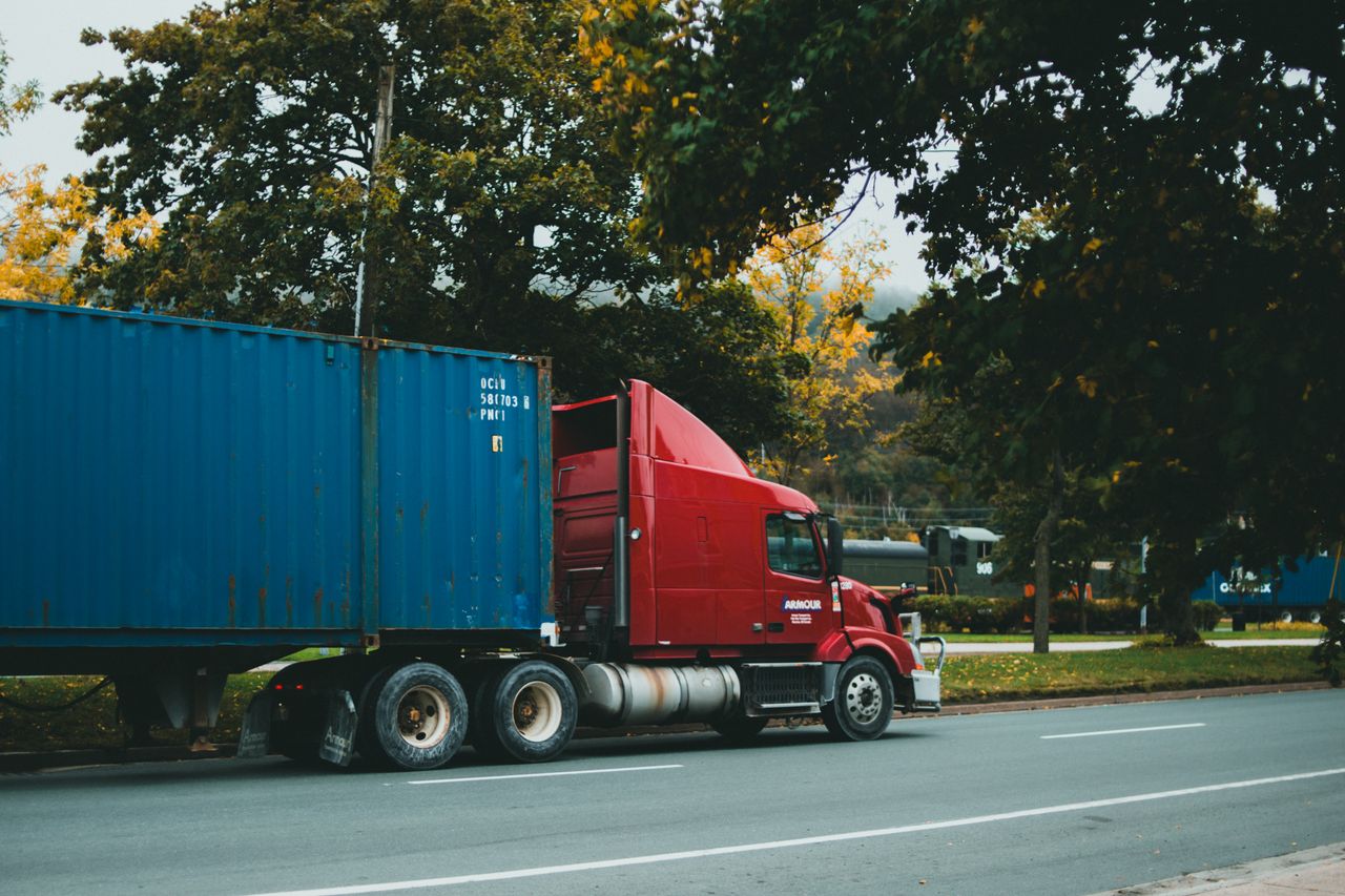 A red truck transports a blue cargo container on a scenic, tree-lined road.